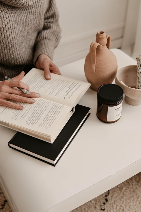 a photo of a person reading a book on a table next to a vase and a candle